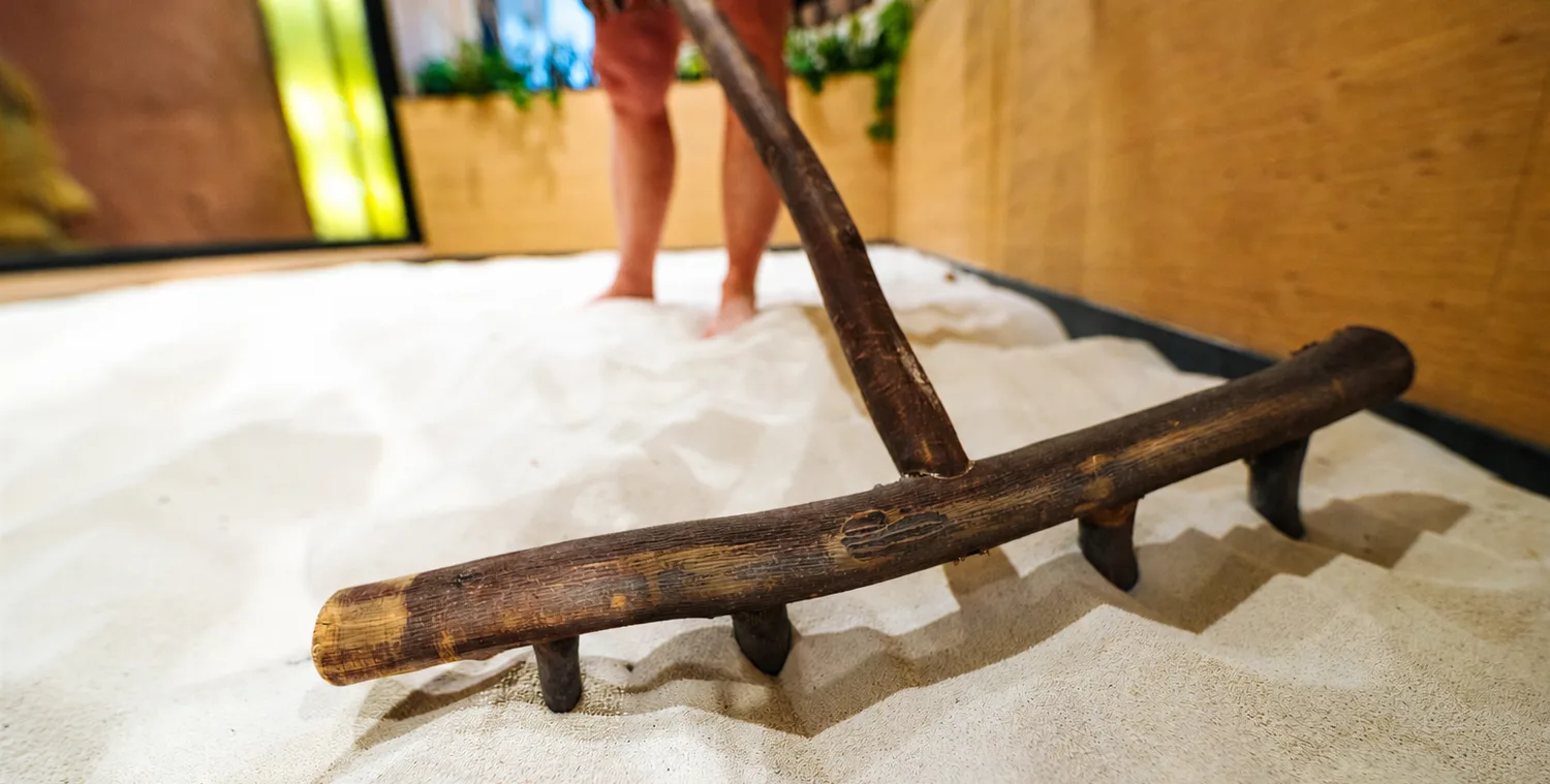 A wooden rake resting in a bed of warm sand while a person stands barefoot in the sand behind it