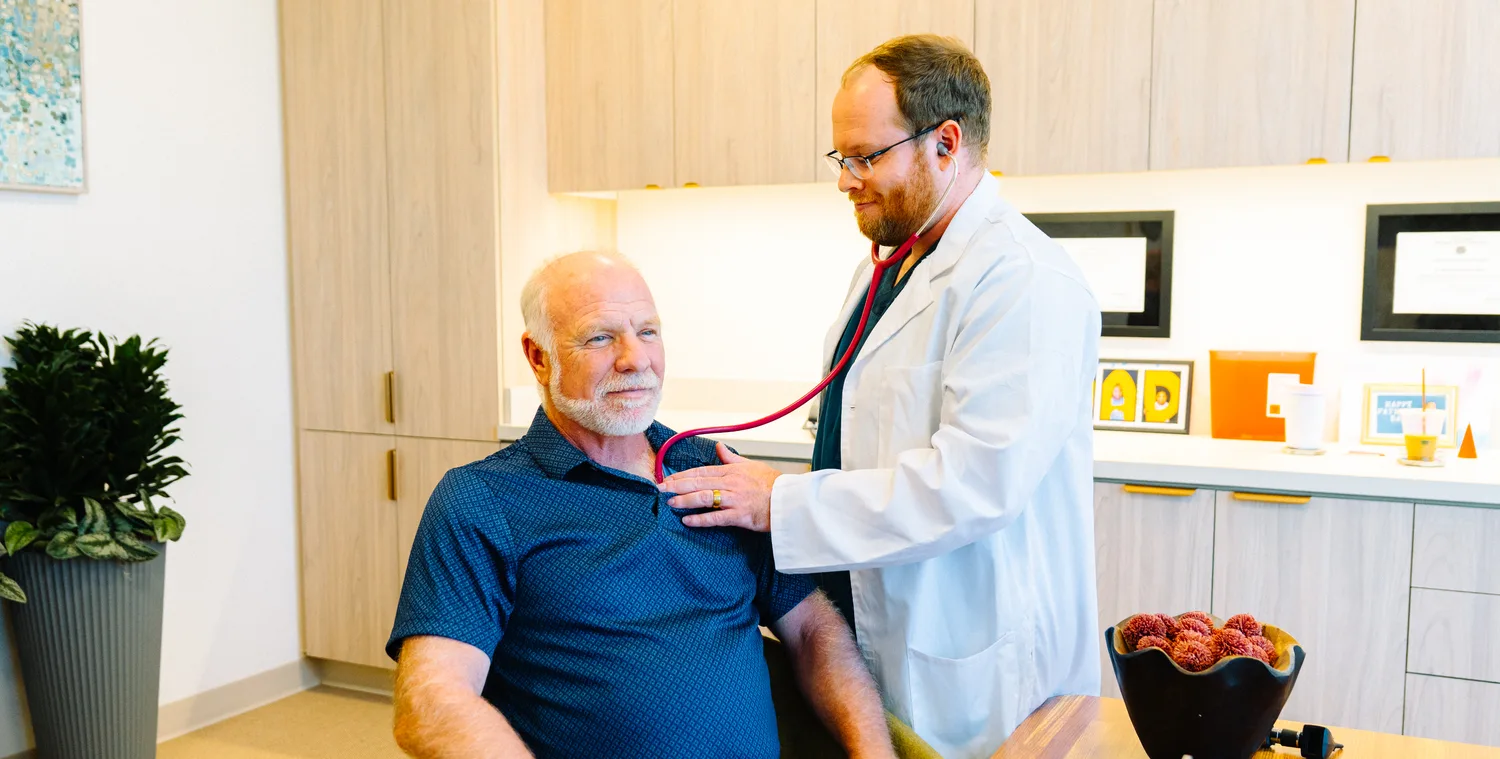 A doctor listening with a stethoscope to an older man’s back during an unhurried check-up in a bright exam room