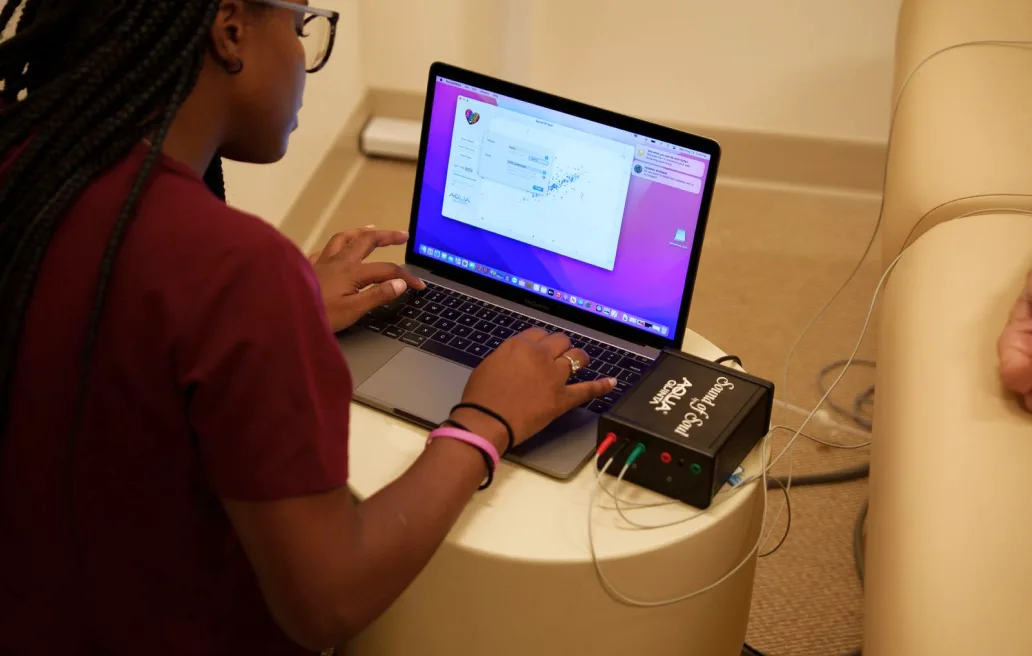 A practitioner in burgundy scrubs, seen from behind, runs an assessment on a laptop wired to a small black diagnostic device on a round side table
