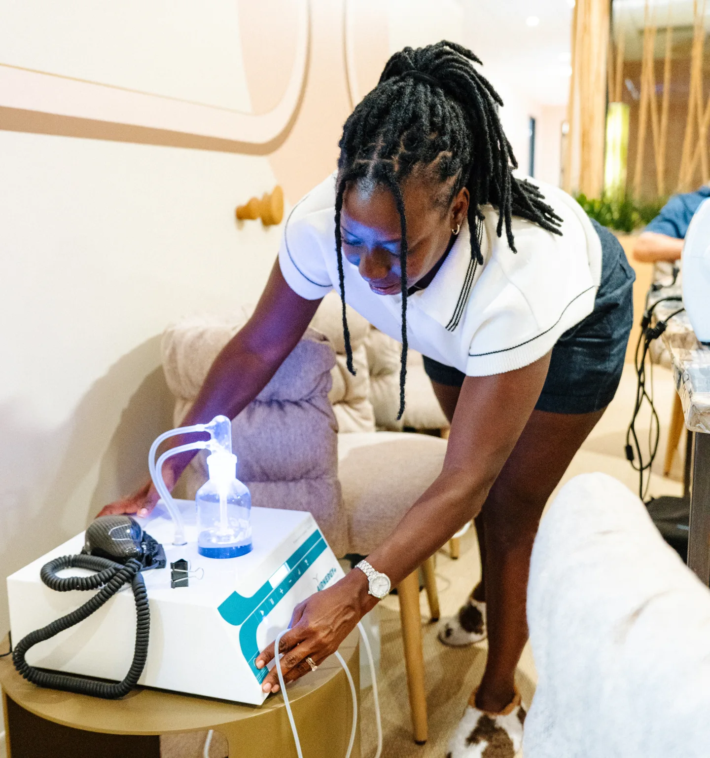 A practitioner in a white polo shirt leans over a white tabletop machine with a water bottle and coiled tubing on a side table in the clinic lounge