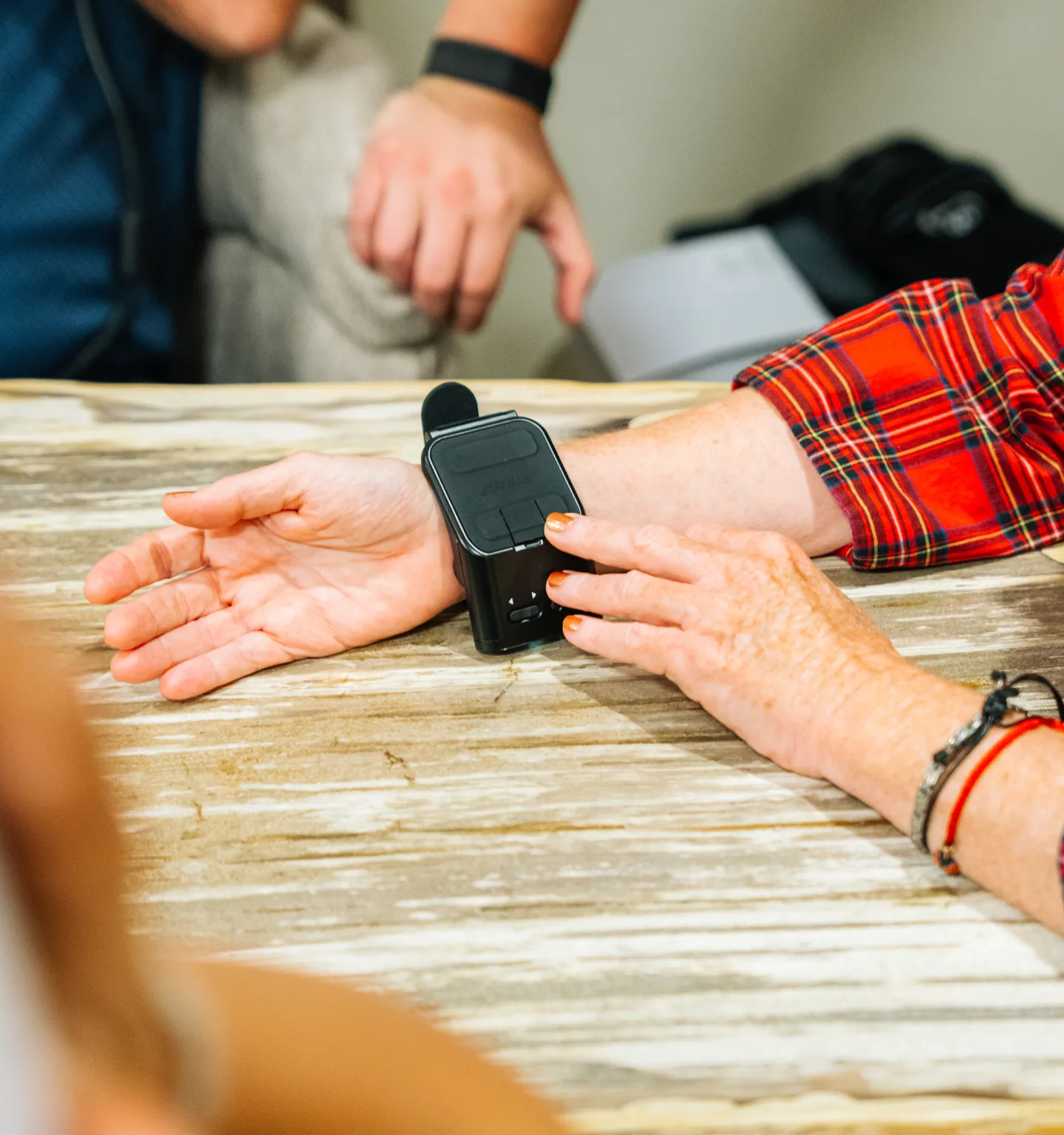 A black laser device worn on a patient’s wrist rests on a marble-look table while another person’s hands steady the arm