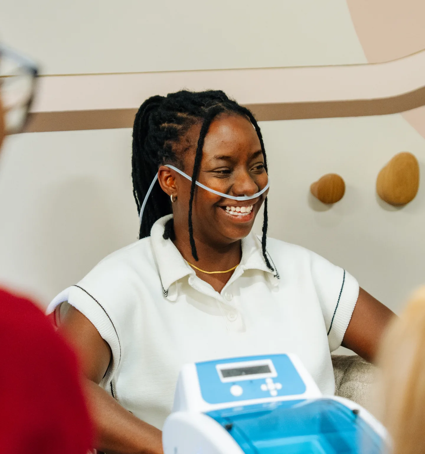 A smiling woman in a white polo shirt breathes through a clear nasal cannula, with a white and blue tabletop device in the foreground