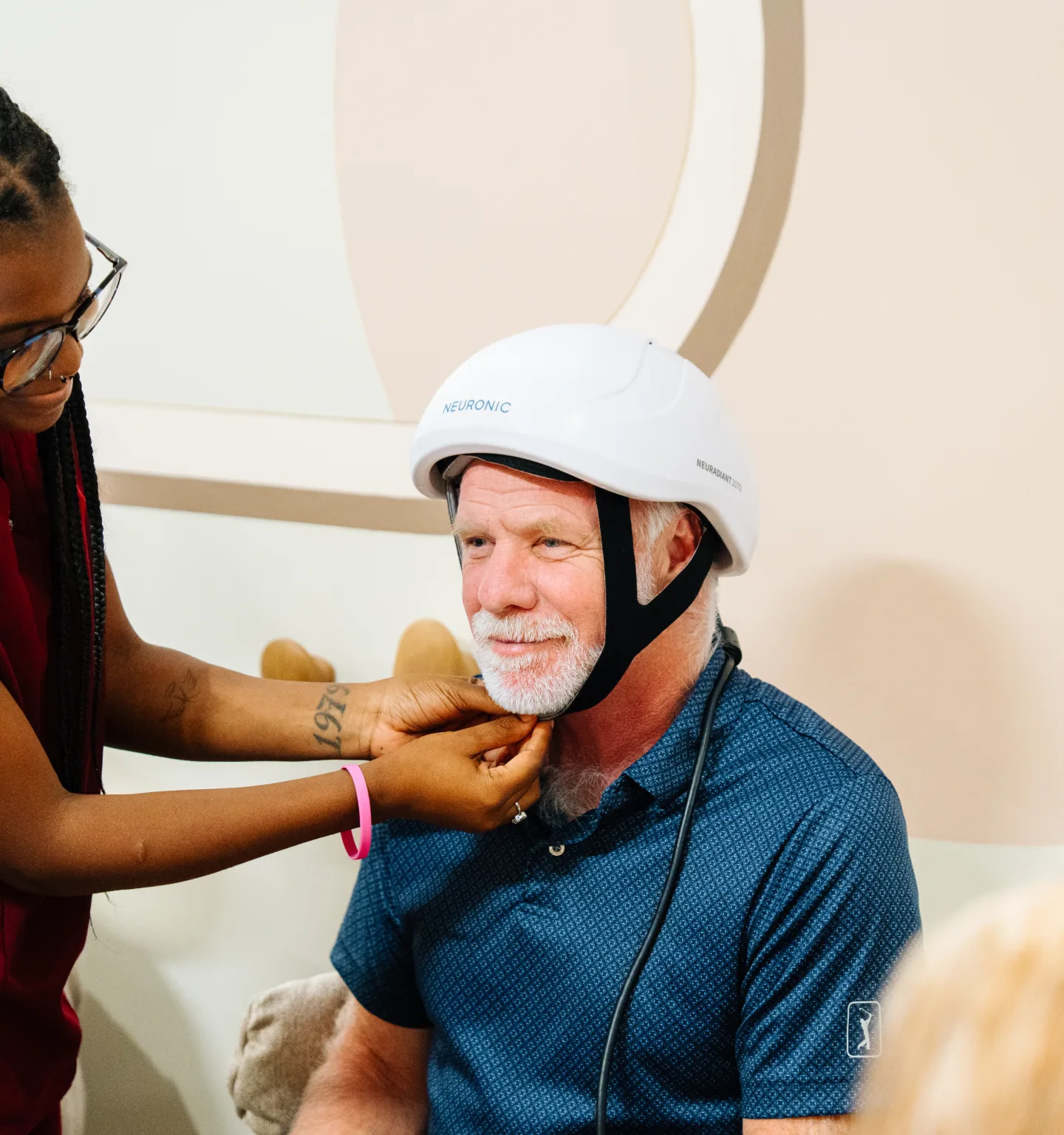 A practitioner fastens the chin strap of a white Neuronic light-therapy helmet on a smiling grey-bearded man in a navy polo shirt