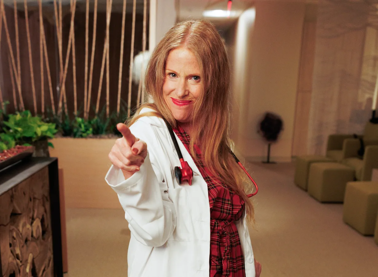 Dr. Sharon Stills, smiling, in a white coat and red plaid dress, points toward the camera in the clinic lounge