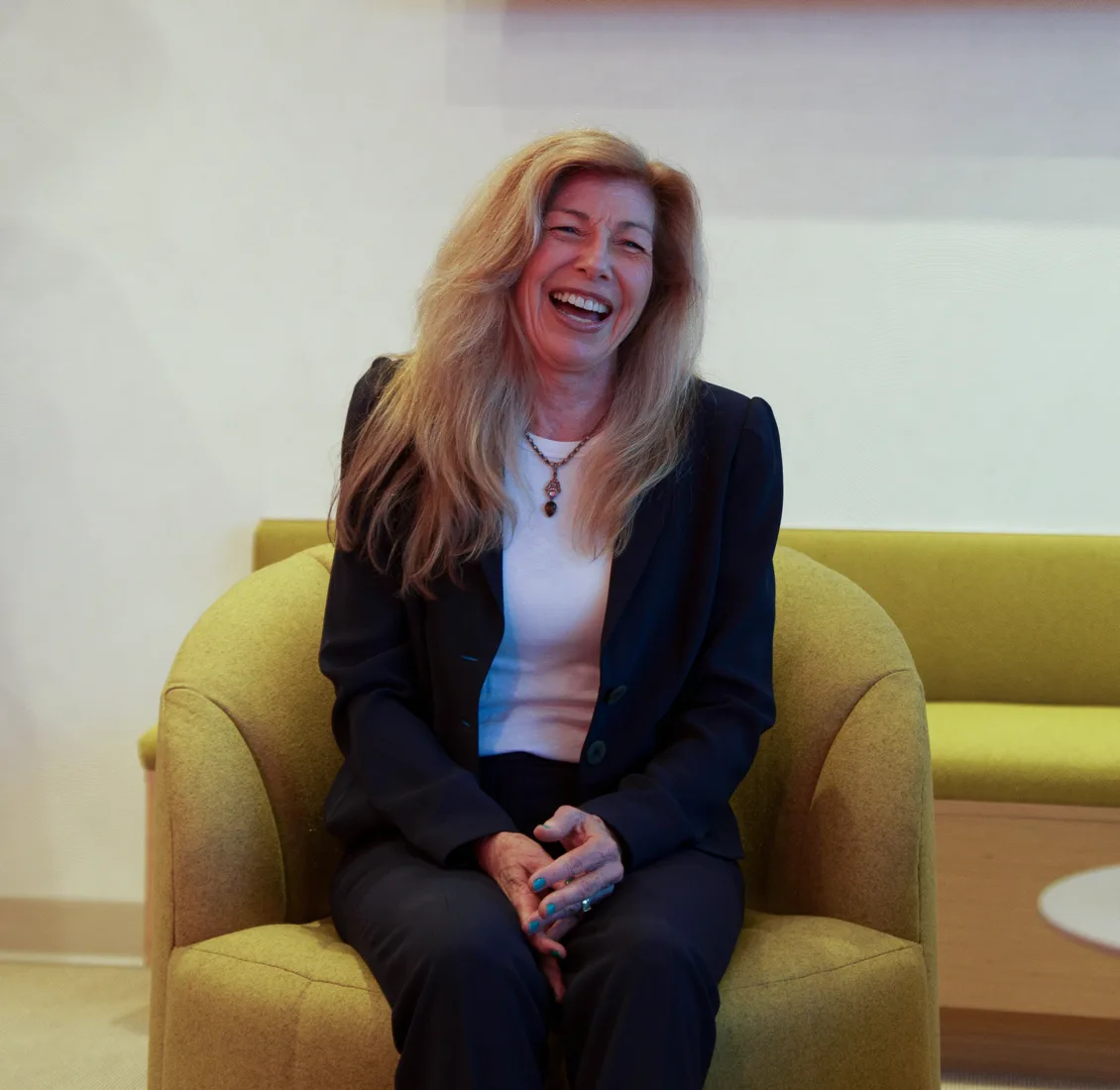 A woman with long blonde hair in a dark blazer laughs, seated in a green armchair in a light waiting area