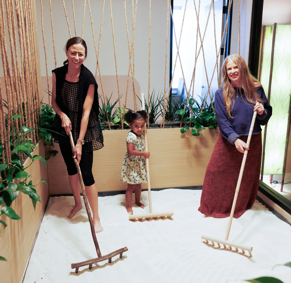 Two women and a small child rake a white sand floor in an indoor garden hung with rope strands and plants