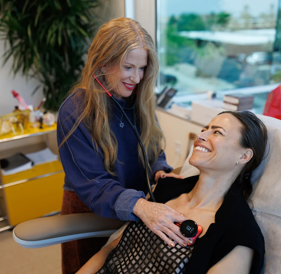 A doctor with long blonde hair holds a small round monitoring device to the chest of a smiling patient reclining in a treatment chair, in a bright clinic room