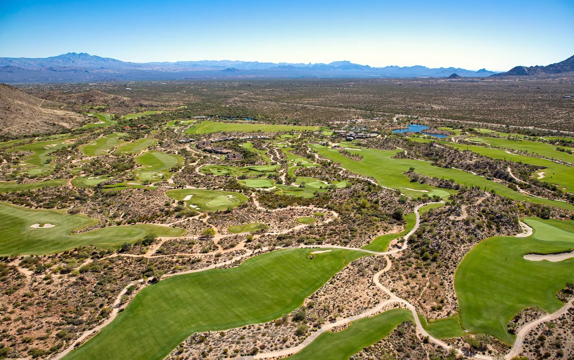 An aerial view of green golf fairways winding through desert scrub, with mountains on the horizon under a clear blue sky