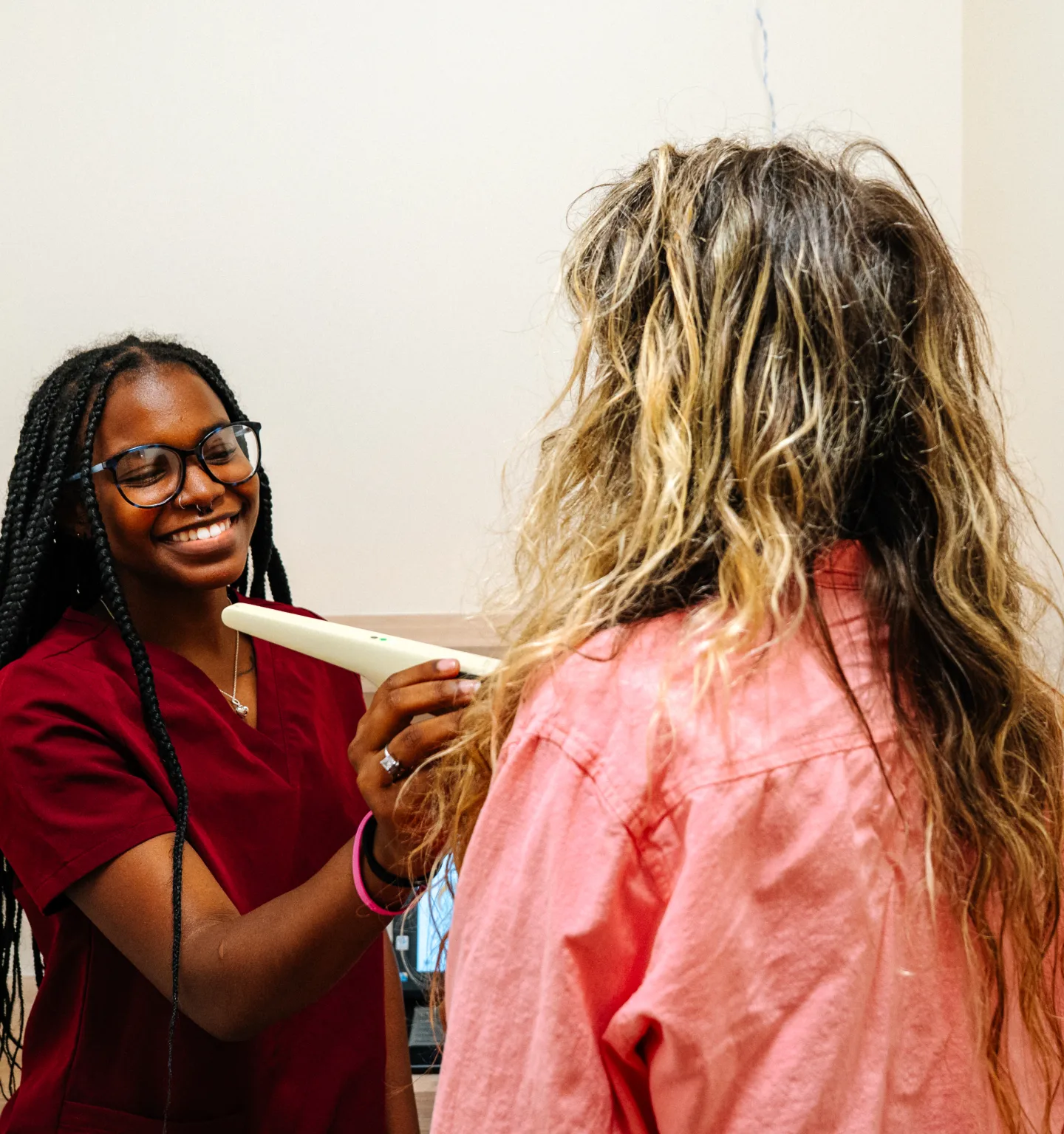 A smiling practitioner in burgundy scrubs and glasses holds a cream-coloured scanning wand to the neck of a woman with long wavy hair in a pink shirt, seen from behind
