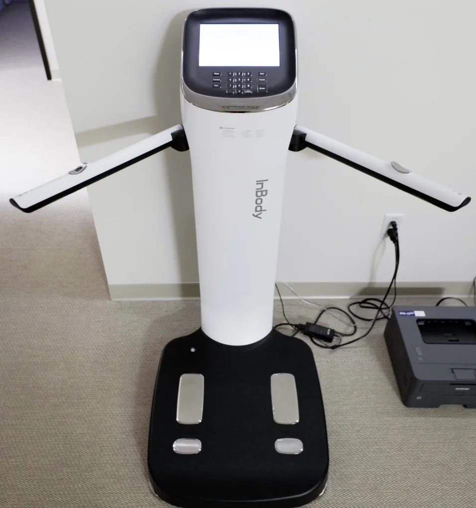 A white InBody body-composition scanner with a touchscreen, two hand grips and a footplate stands on grey carpet beside a printer