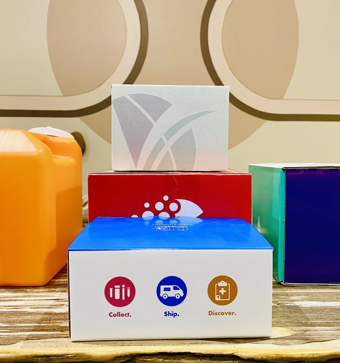 Lab test-kit boxes in white, red, blue and purple are stacked beside an orange container on a wooden counter; the front box reads “Collect. Ship. Discover.”