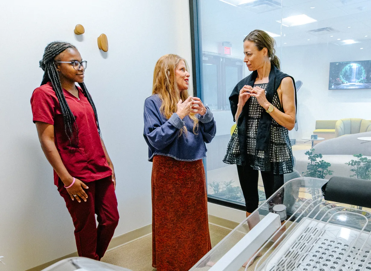 Three women talk in a bright treatment room, one in maroon scrubs leaning on the wall, one red-haired in a navy sweater