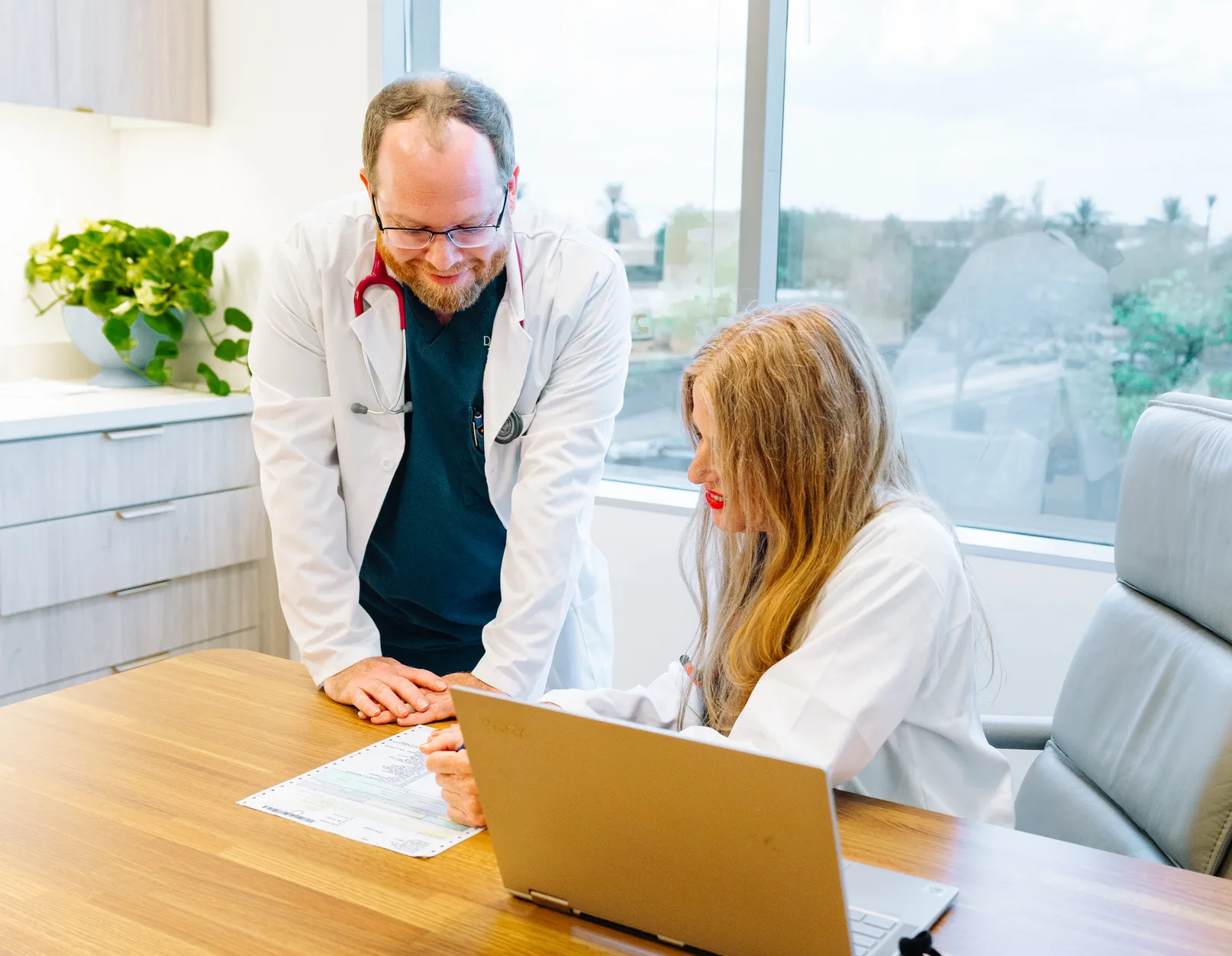 Two physicians in white coats at an office desk, reviewing papers beside a laptop