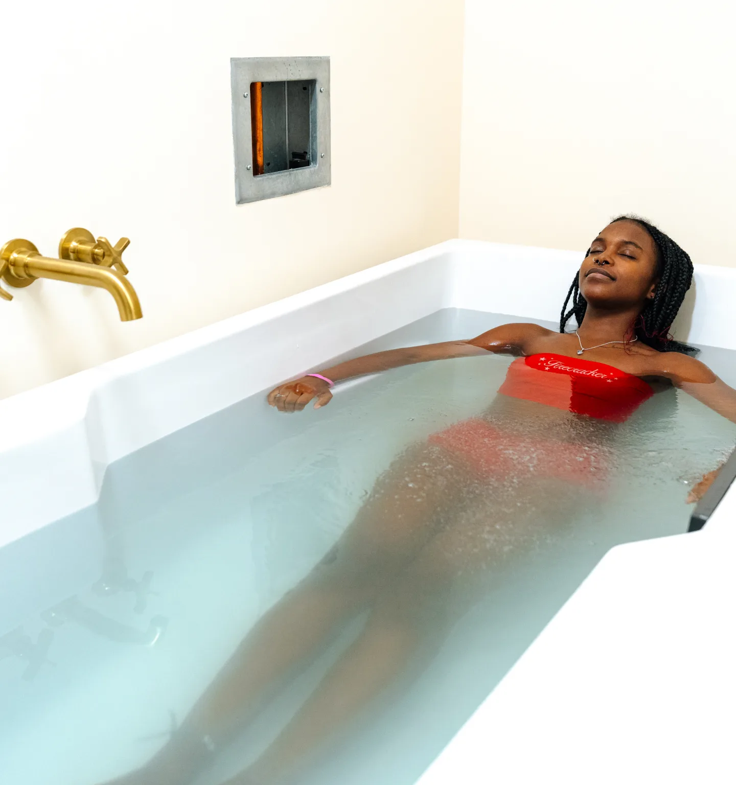 A young woman in a red swimsuit relaxes with her eyes closed in a white bathtub of milky, oxygen-rich water beneath a brass wall tap