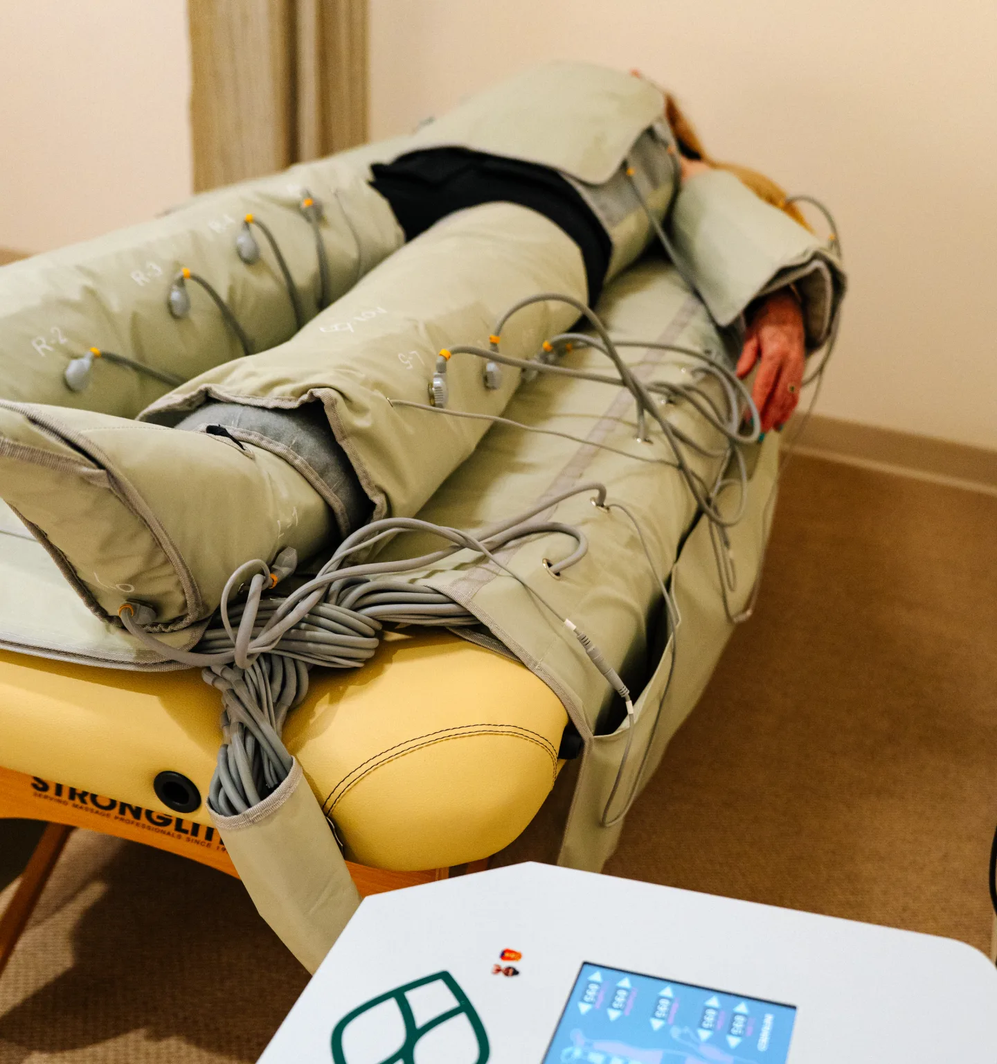 A patient lies on a padded yellow table wrapped from the legs up in grey pneumatic compression garments, with the control unit's touchscreen in the foreground
