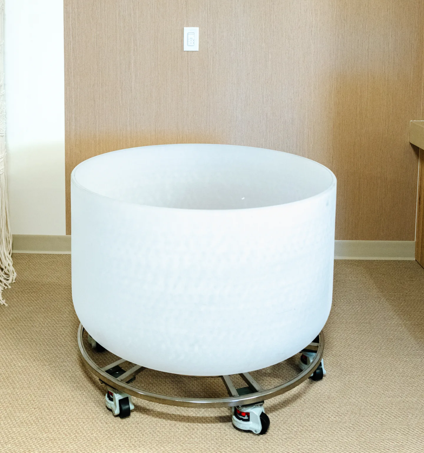 A large frosted-white crystal singing bowl on a wheeled steel stand in a quiet room with beige walls and carpet