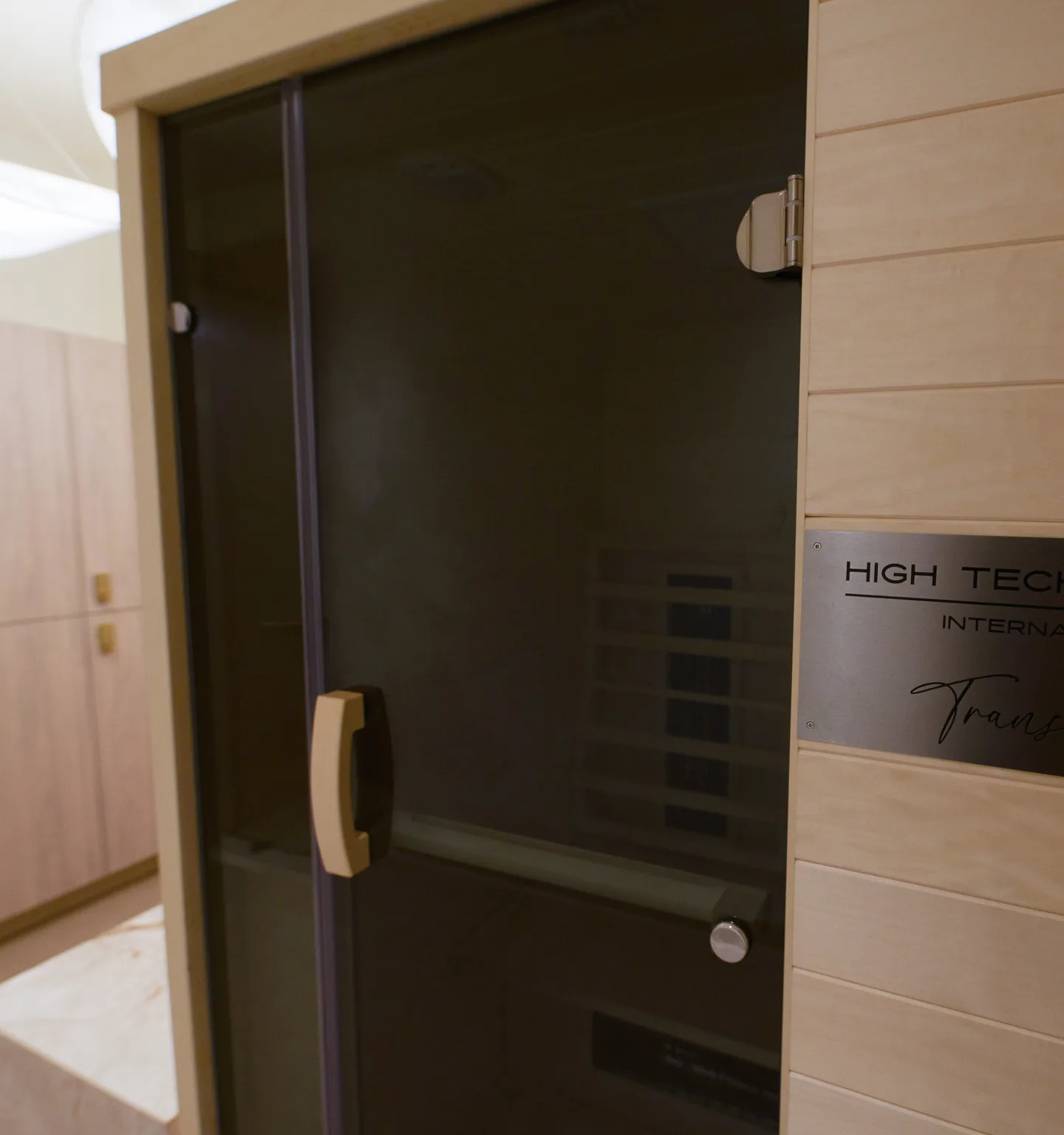 A pale-wood infrared sauna cabin with a closed smoked-glass door and a brushed-steel maker's plaque on its side panel