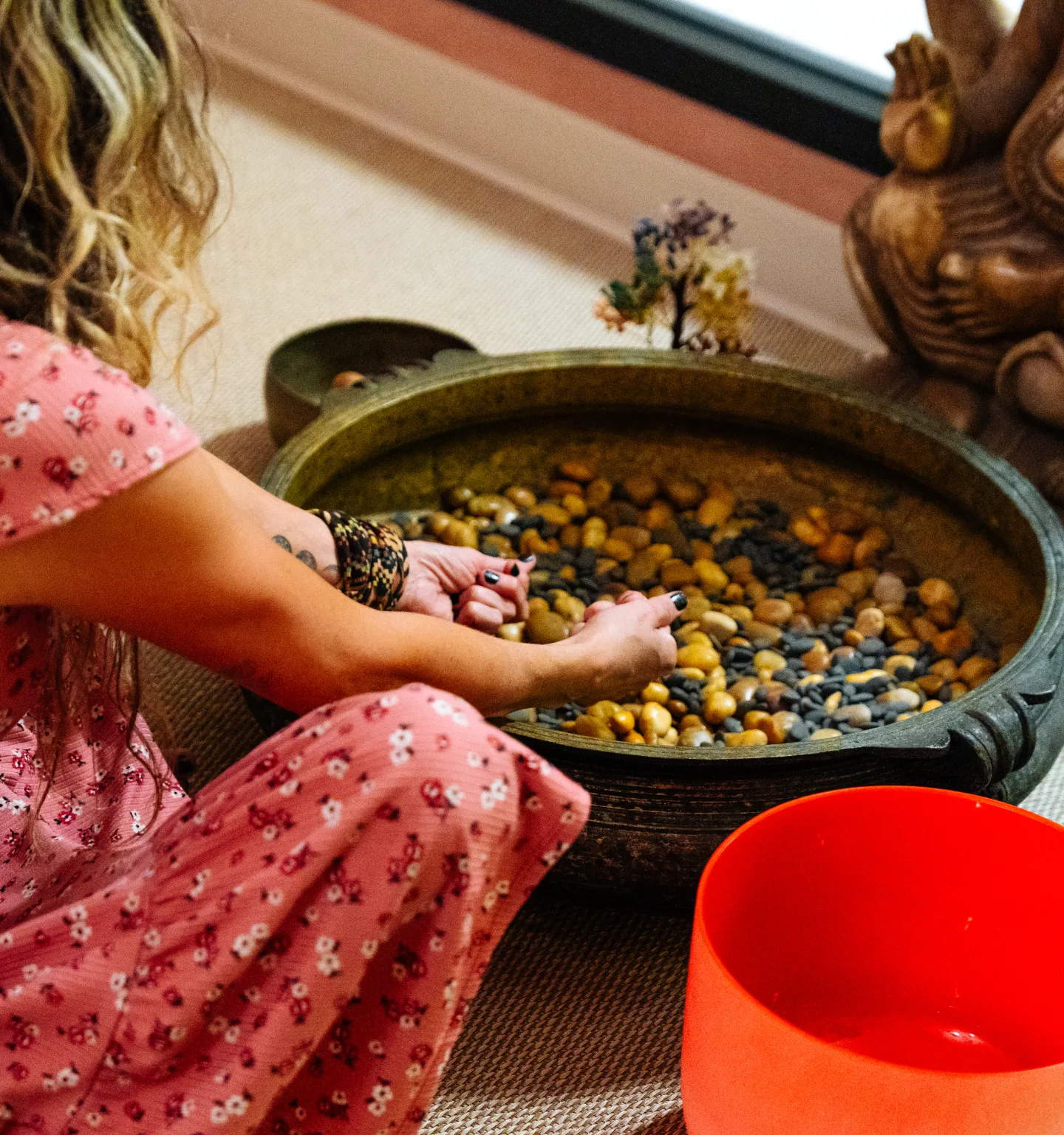 A woman in a pink floral dress runs her hands through polished stones in a large brass bowl, beside a carved statue, a small gemstone tree and an orange crystal singing bowl