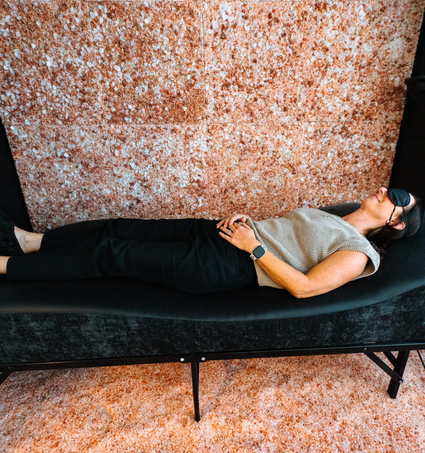 A woman wearing an eye mask lies on a black vibroacoustic bed in front of a pink Himalayan salt wall, with salt covering the floor