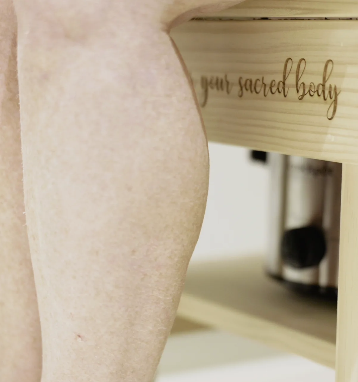 A close-up of a bare leg beside a pale wooden steaming seat engraved "your sacred body", with the steam pot visible beneath it