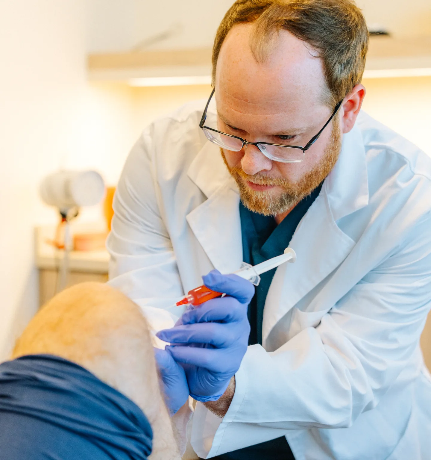 A bearded doctor in glasses and a white coat, wearing purple gloves, leans in to give an injection with an orange-capped syringe at a patient's knee