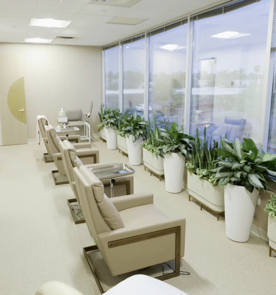 A bright IV lounge with a row of beige recliners and side trays along floor-to-ceiling windows lined with leafy plants in tall white planters