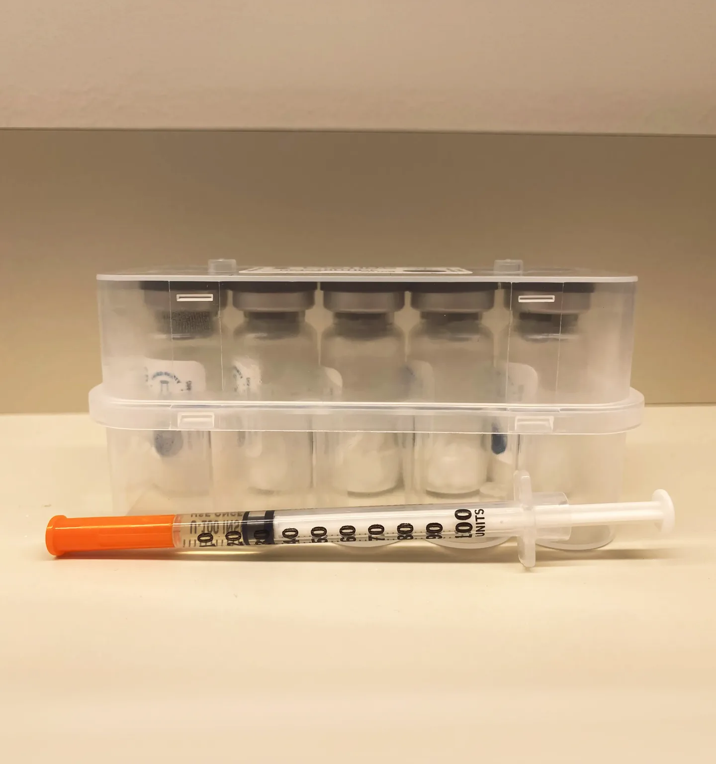 A clear plastic case holding five small vials stands on a cream counter, with an orange-capped insulin syringe lying in front of it
