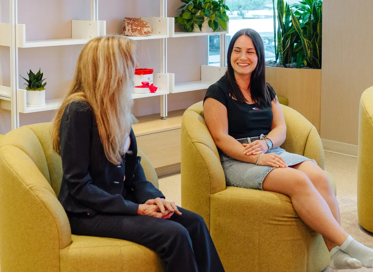 Two women talk in yellow armchairs in the clinic lounge; one, smiling, faces the camera