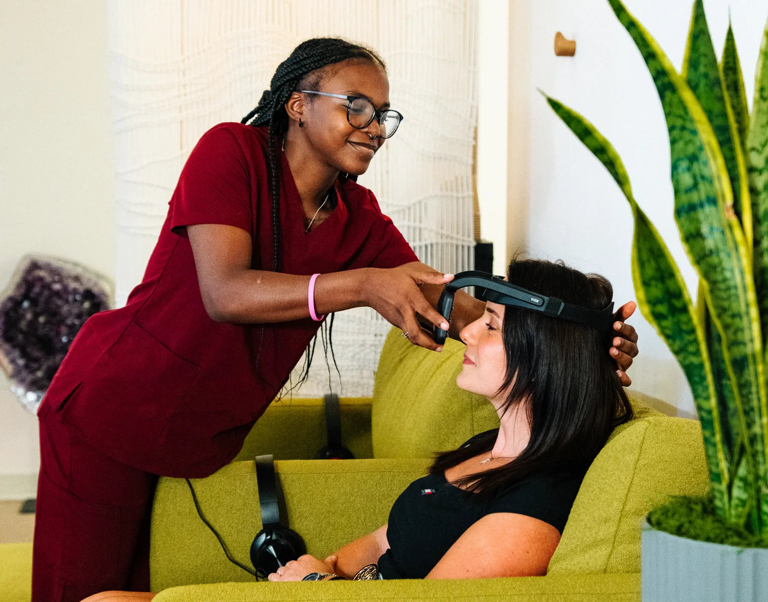 A practitioner in red scrubs fits a headset on a woman reclining on a green sofa
