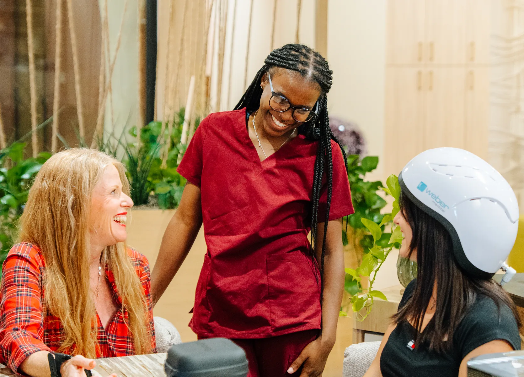 A practitioner in red scrubs laughs with two seated women, one wearing a light-therapy helmet