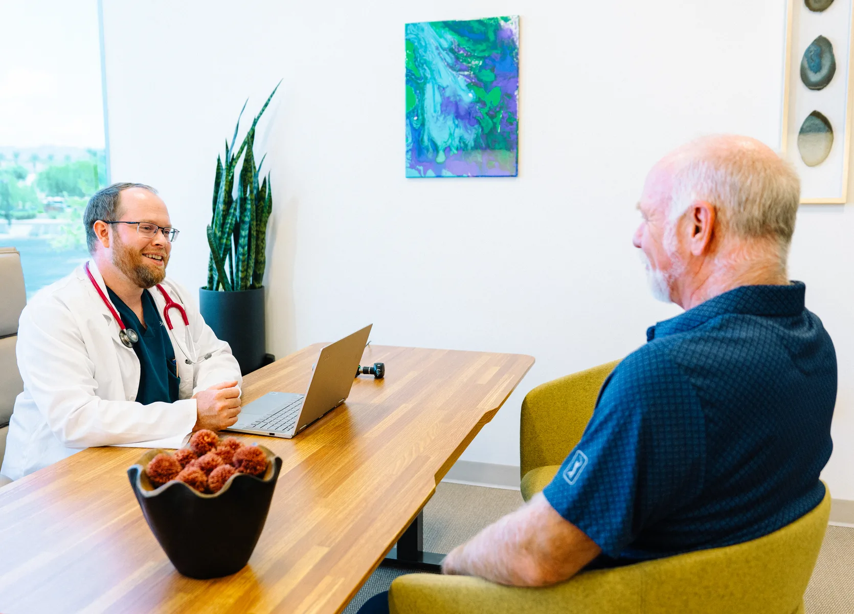 A doctor in a white coat talks with an older man across a wooden table