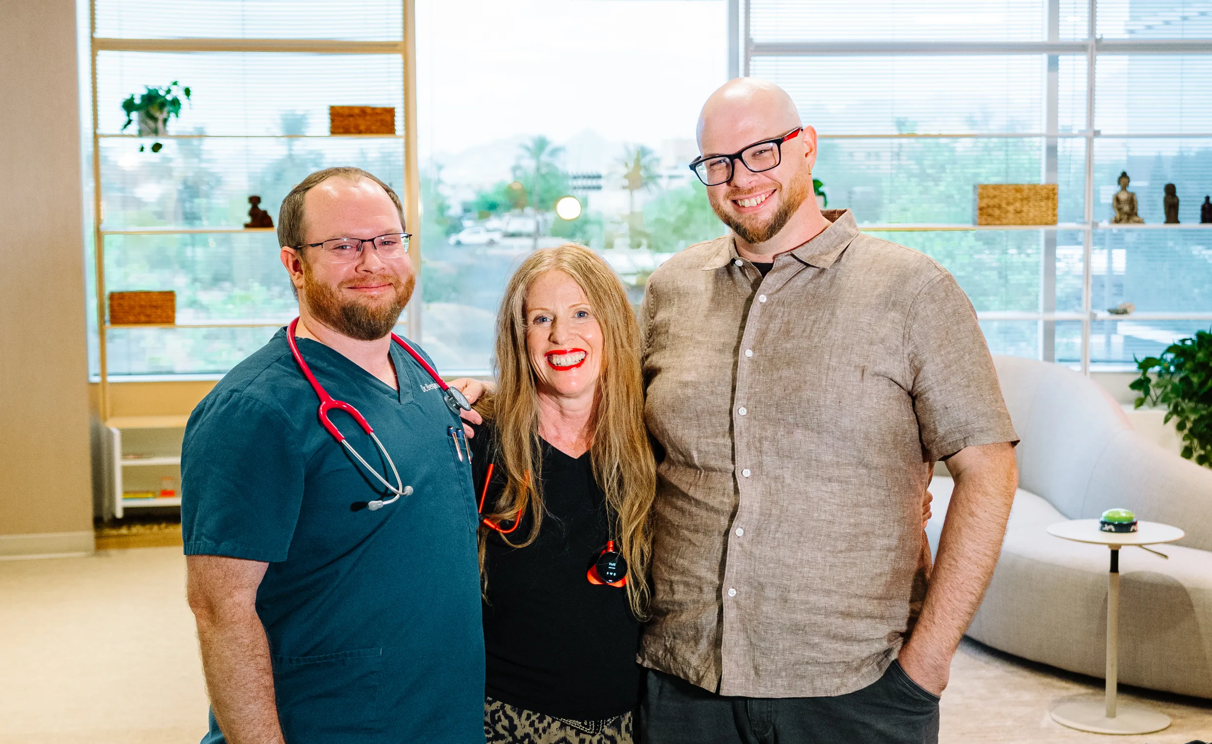 Three smiling people stand arm in arm in the bright clinic lounge: a man in teal scrubs with a stethoscope, a woman with long red hair, and a bald man in glasses