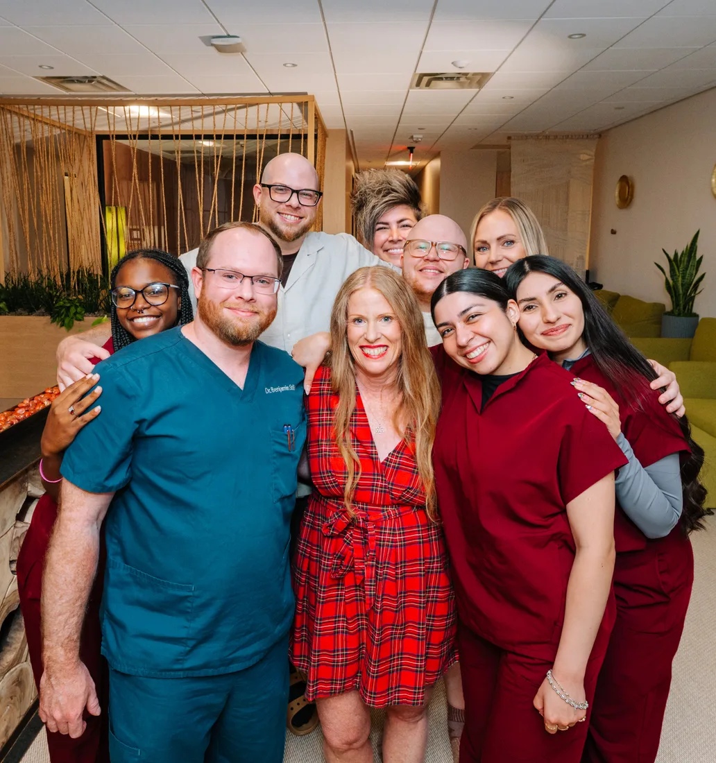 The clinic's team, nine smiling staff members in teal and burgundy scrubs, a white tunic and a red plaid dress, hugging together in a lounge with rope-curtain partitions and olive-green sofas