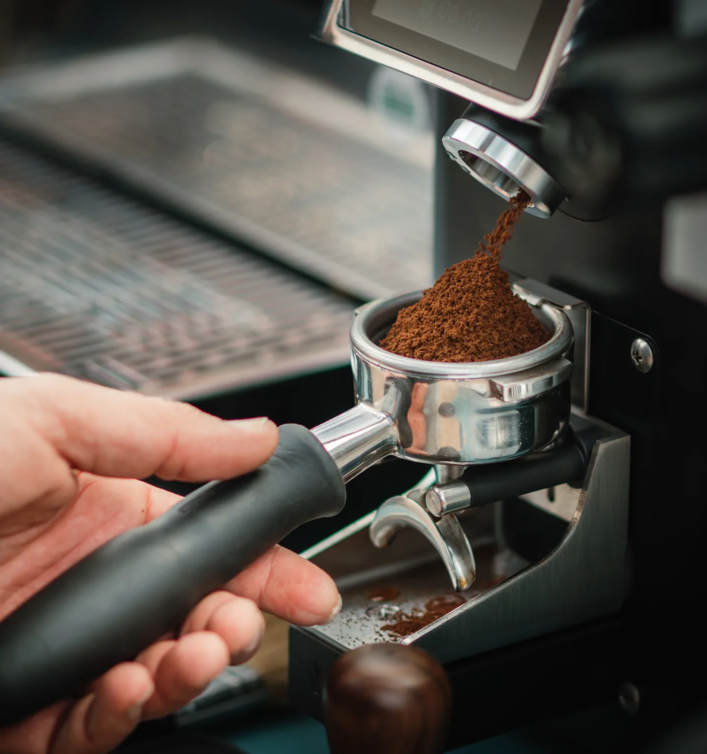 A close-up of a hand holding an espresso portafilter as freshly ground coffee falls into it from a black grinder