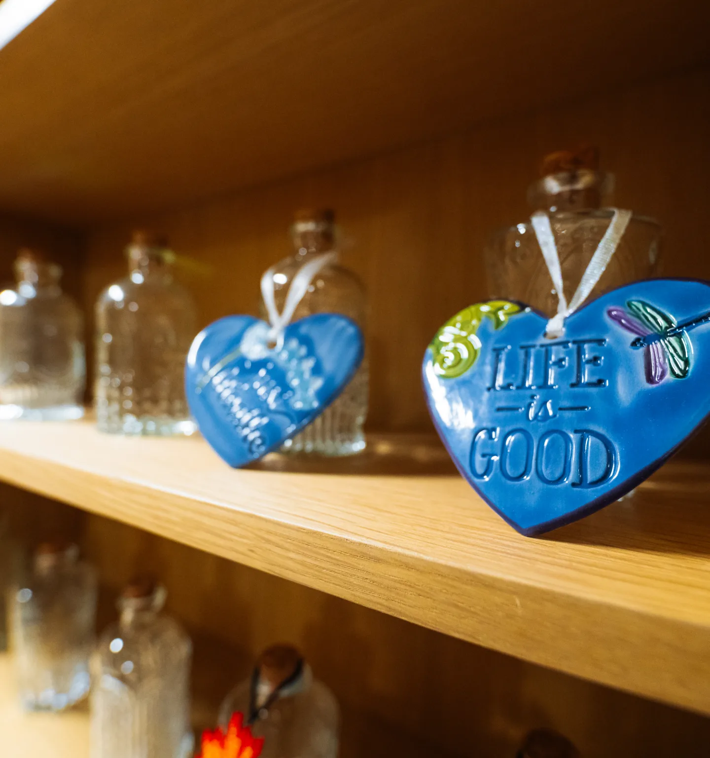 Two blue ceramic heart ornaments hang from glass bottles on a pale wooden shelf; the nearer one reads "Life is Good" beside a painted dragonfly