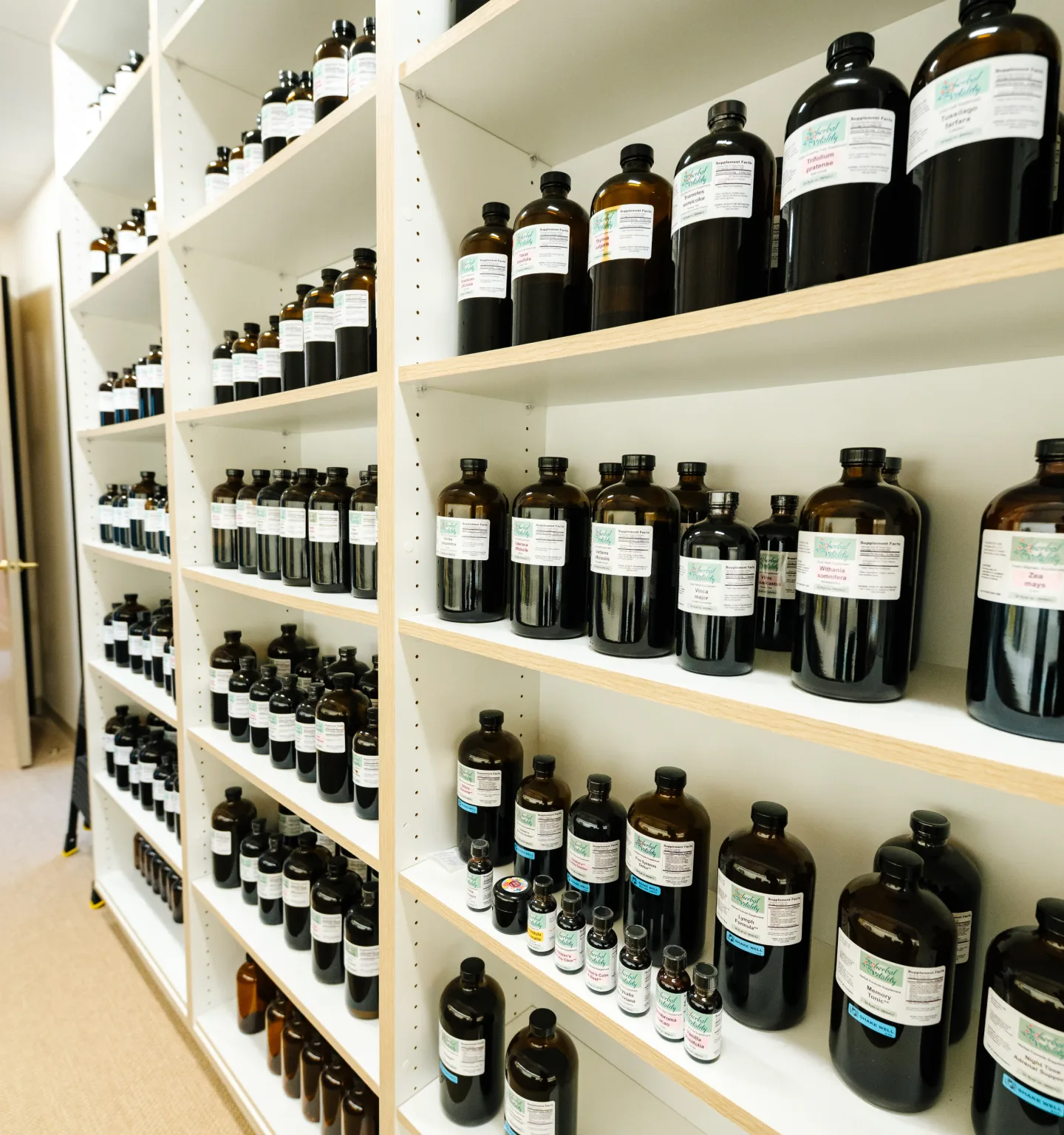 Floor-to-ceiling white shelves lined with rows of labelled amber glass tincture bottles along a bright corridor