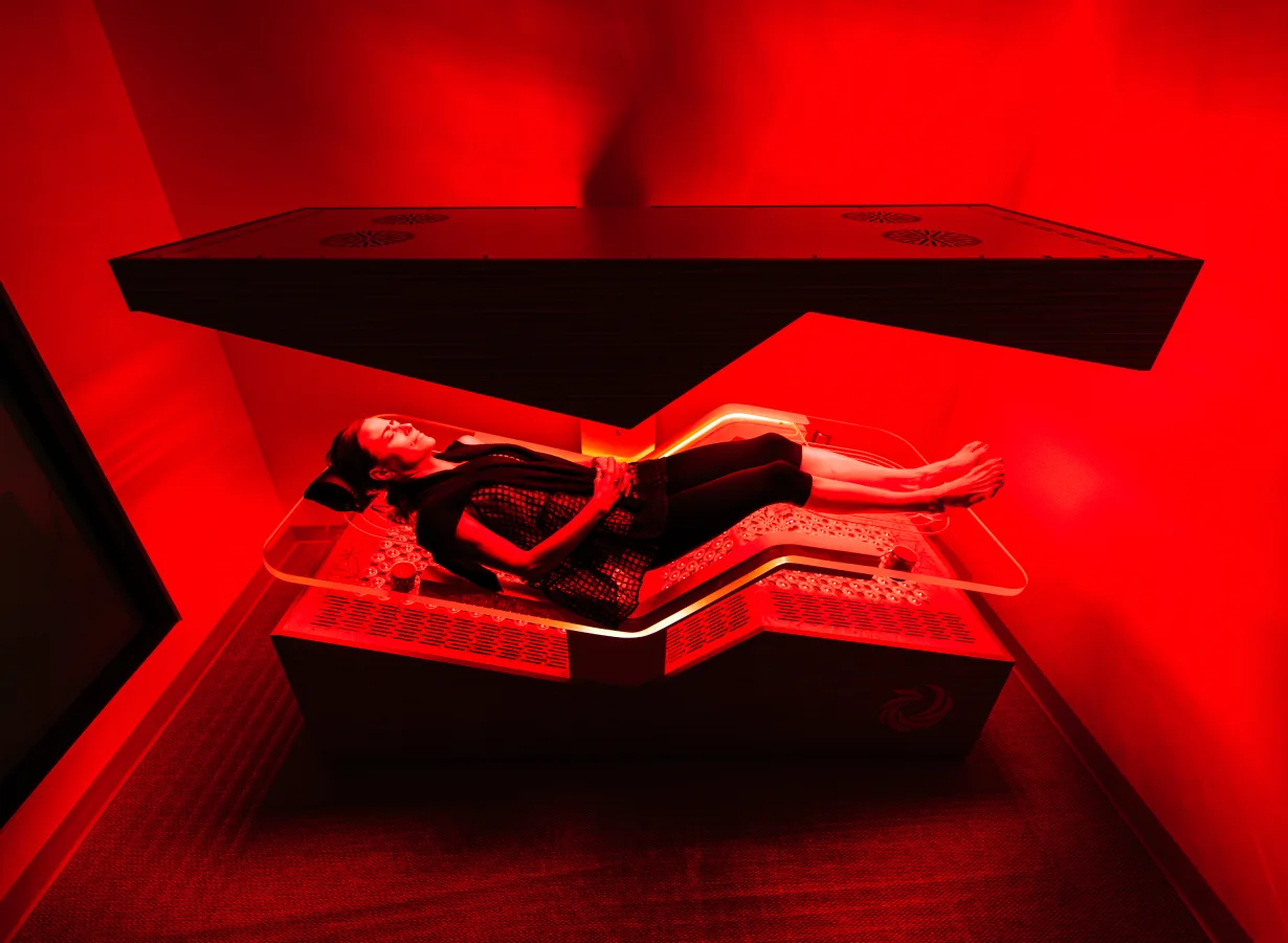 A woman reclines on a glowing red-light therapy bed beneath a matching suspended panel, the treatment room bathed in red light