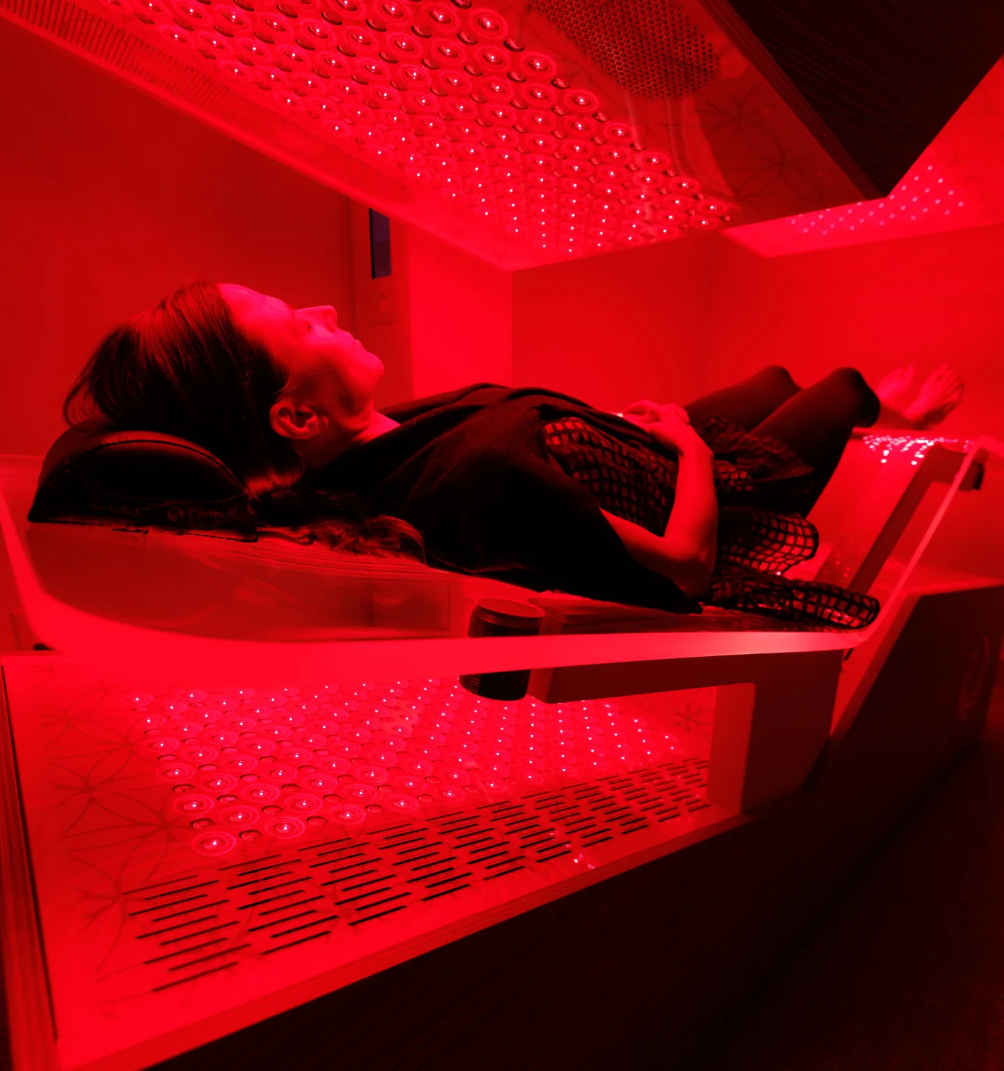 A woman lies on her back on the padded bed of a regenerative chamber, bathed in red light from panels above and below her