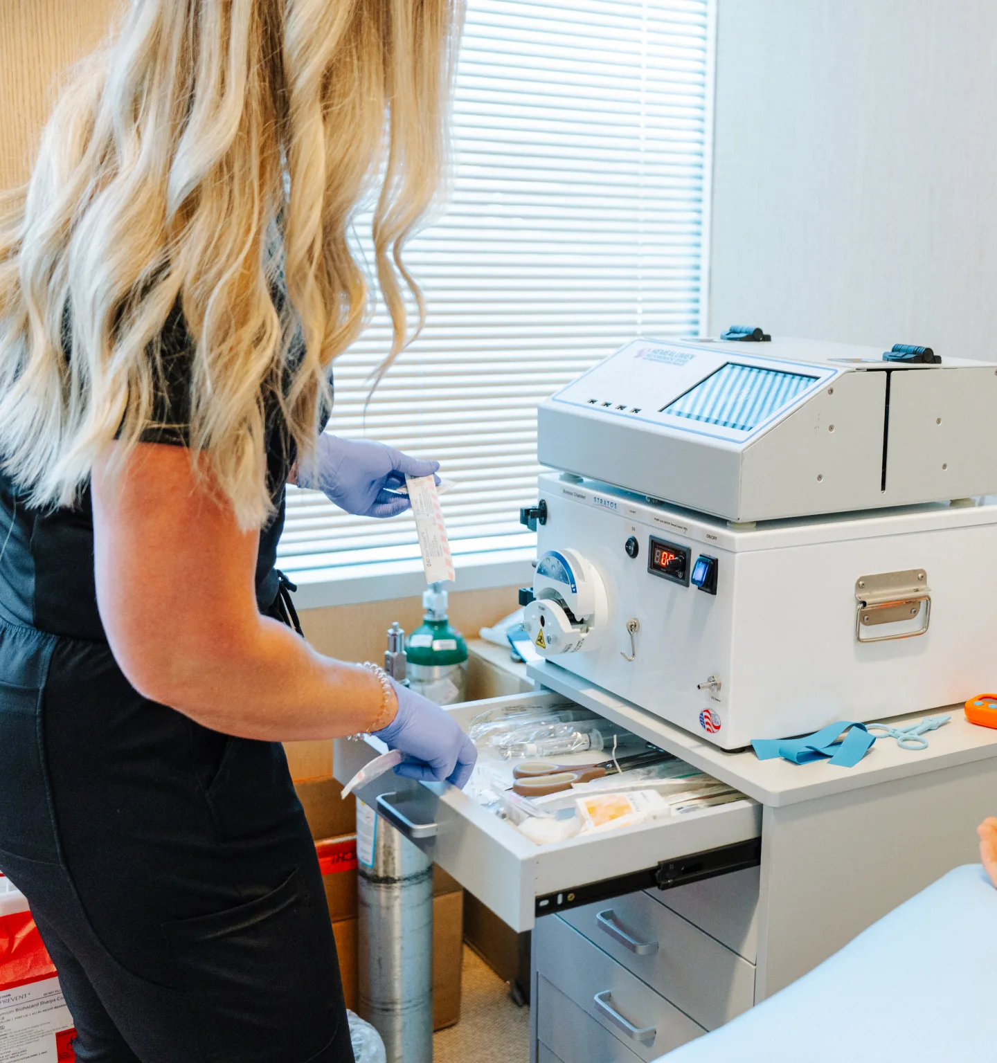 A practitioner with long wavy blonde hair, in black scrubs and blue gloves, prepares supplies at a white blood-filtration machine on a wheeled cart beside a window with blinds