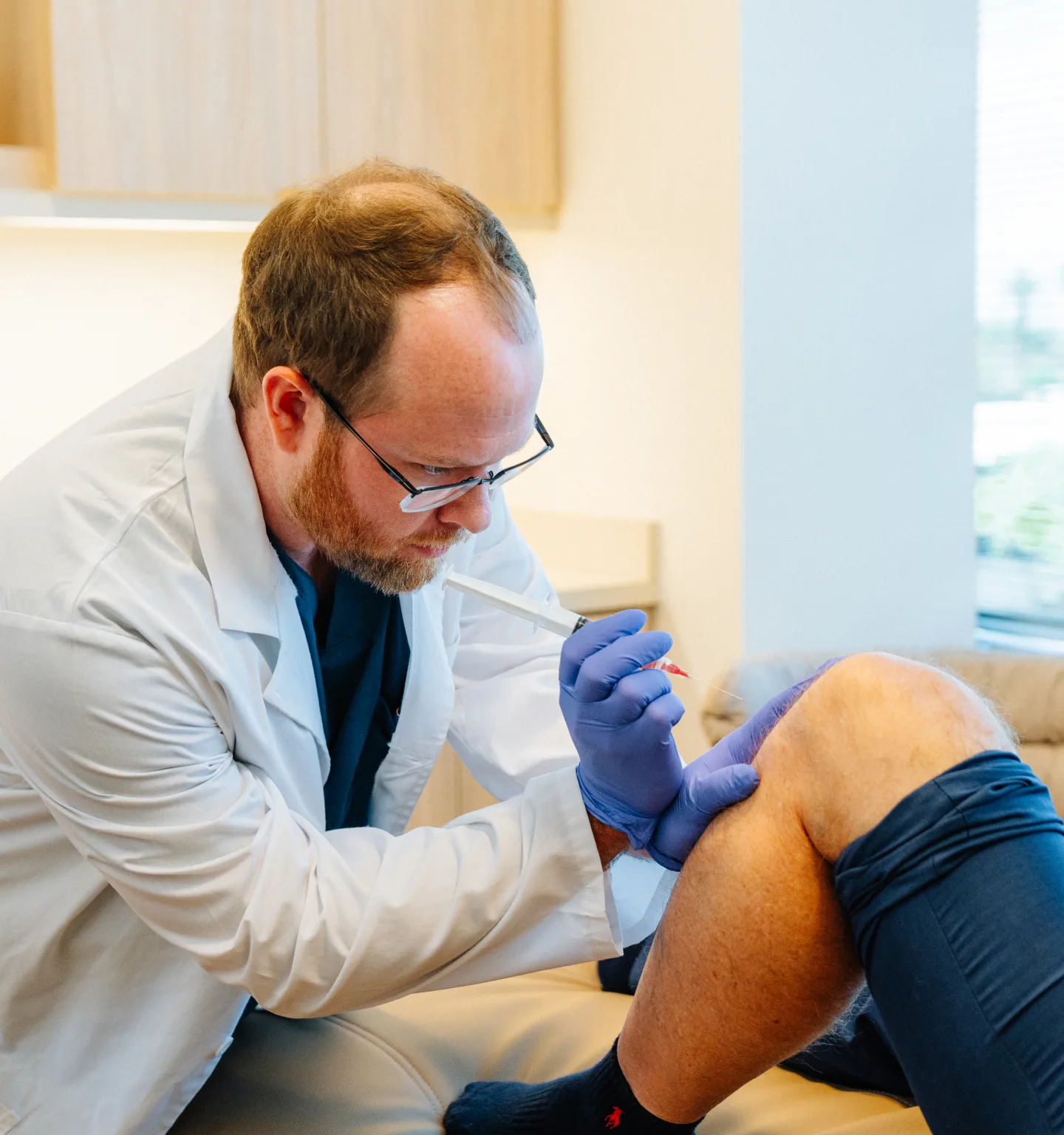 A bearded doctor in glasses and a white coat, wearing purple gloves, gives an injection into the knee of a seated patient in dark shorts